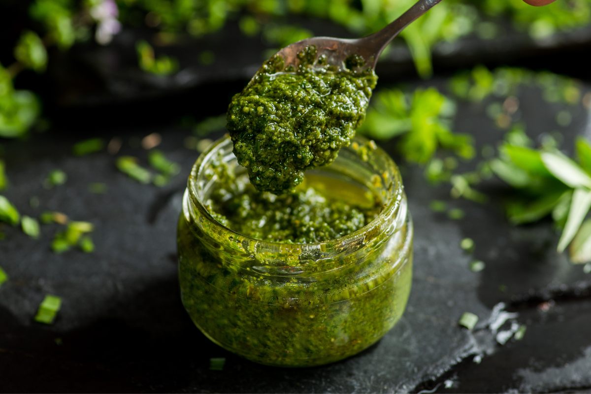 Spoon lifting vibrant green pesto from a jar, surrounded by scattered fresh herbs on a dark surface.