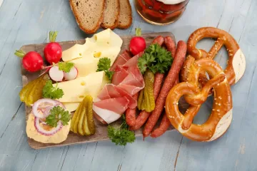 Wooden board of Bavarian cold cuts — Emmental cheese, cured ham, thin smoked sausages, gherkins, radishes and Obatzda — with two pretzels, rye bread and a beer alongside