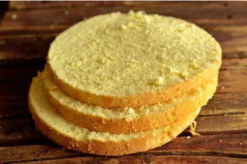 Three slices of a pale, fine-crumbed gluten-free sponge cake stacked on a rustic wooden board