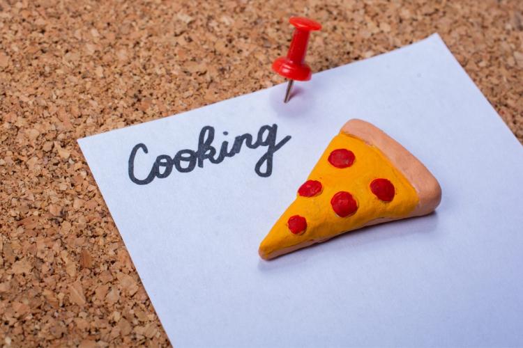 A note pinned to a corkboard with the word 'Cooking' written on it and a small clay pizza slice attached.