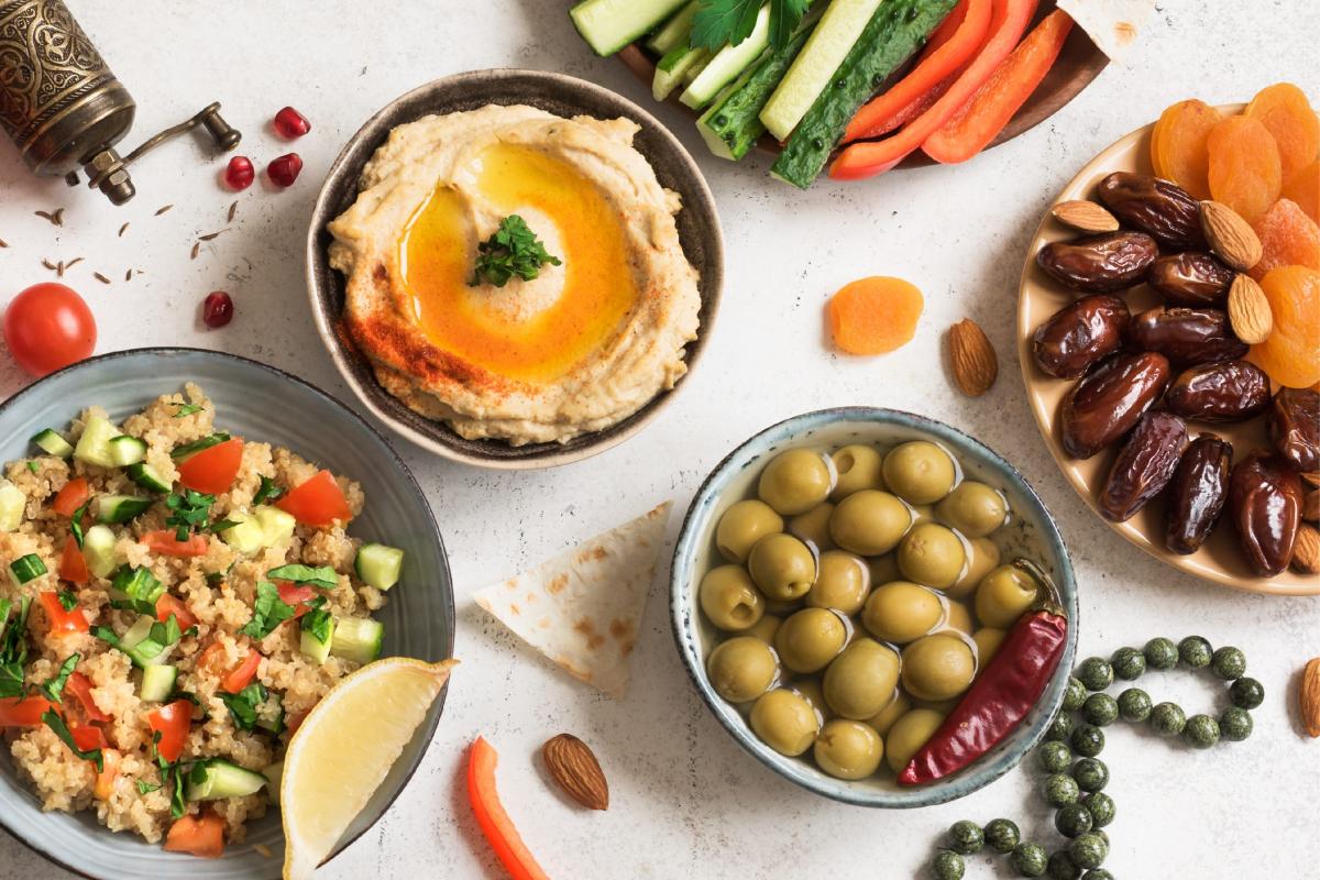 Middle Eastern food spread with a bowl of hummus topped with olive oil and paprika, fresh vegetable sticks, green olives with a red chili pepper, couscous salad with cucumber and tomato, and a plate of dried fruits with almonds. Garnishes include a vintage pepper grinder, cherry tomatoes, and a lemon wedge.
