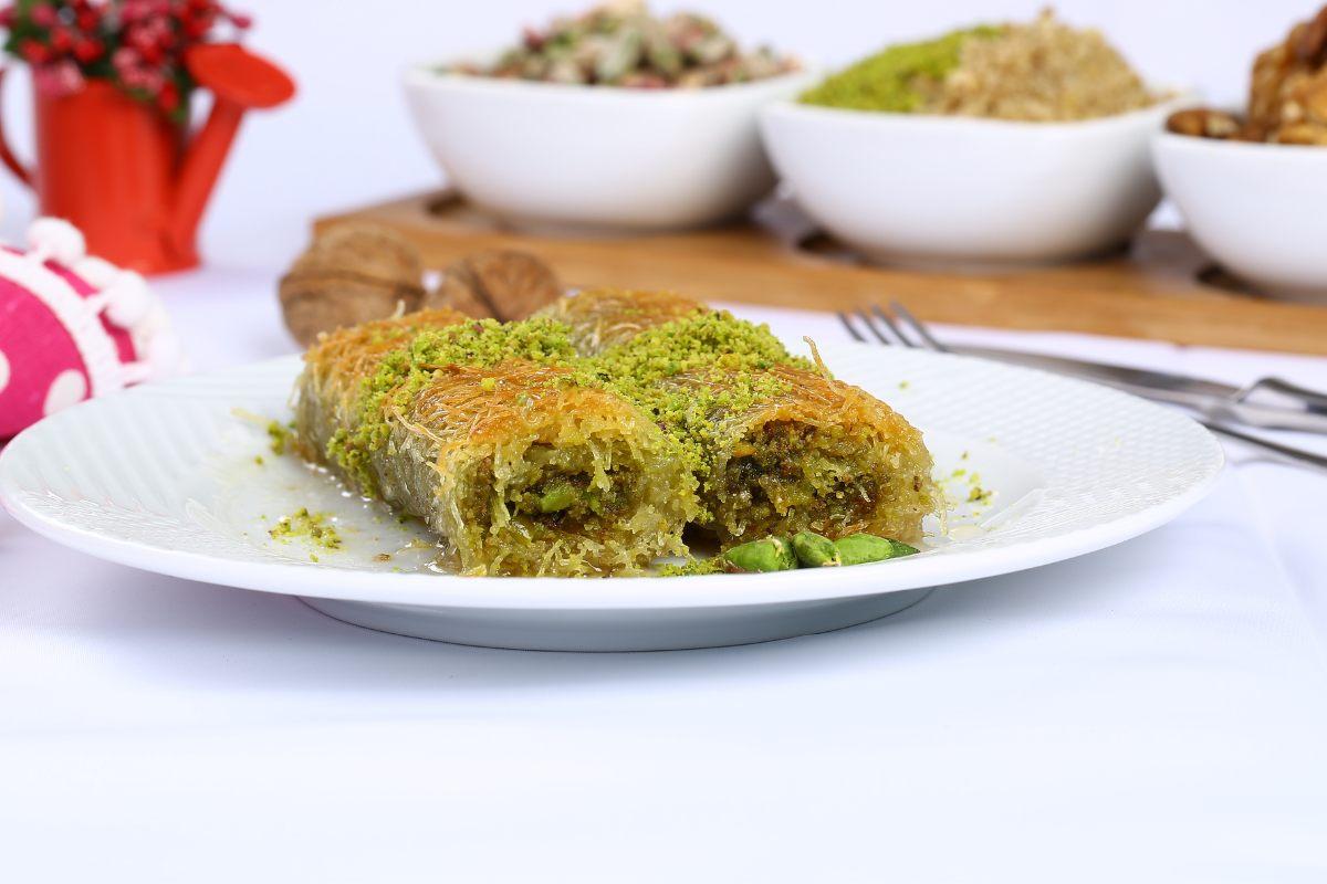Two pieces of rolled kataifi on a white plate, decorated with chopped pistachios, bowls of ingredients at the background.