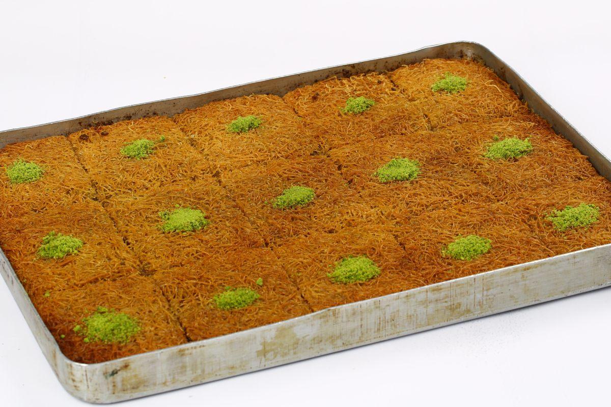 A rectangular baking tray filled with baked kataifi cut into squares, each topped with chopped pistachios.