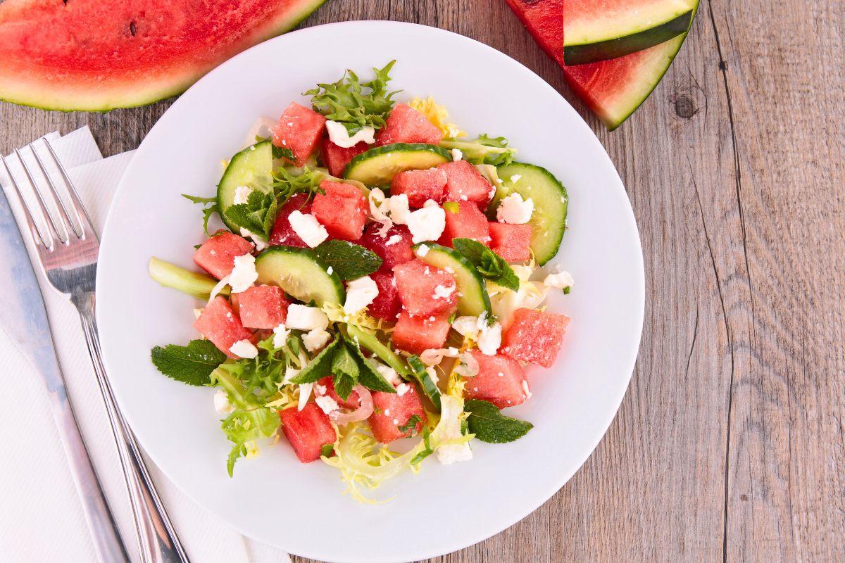 A colorful watermelon and feta salad with watermelon cubes, cucumber slices, crumbled feta cheese, fresh mint, and mixed greens on a white plate, placed on a rustic wooden table.