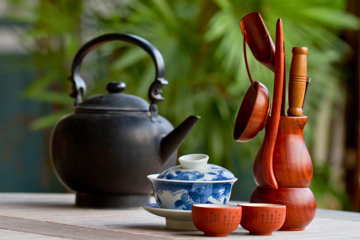 A traditional Chinese tea set with a black teapot, blue and white porcelain gaiwan, reddish-brown clay teacups, and wooden tea utensils arranged on a table against a green, natural background.