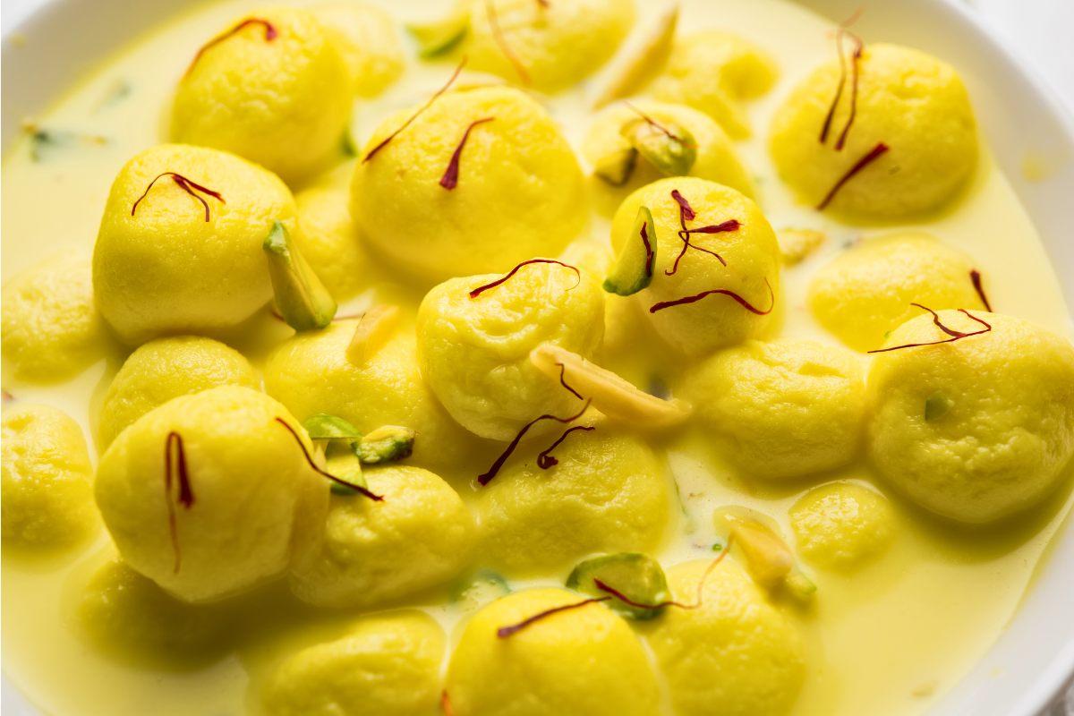 Close-up of Rasmalai, featuring yellow paneer balls soaked in saffron-infused milk, garnished with slivers of pistachios, almonds, and saffron strands.