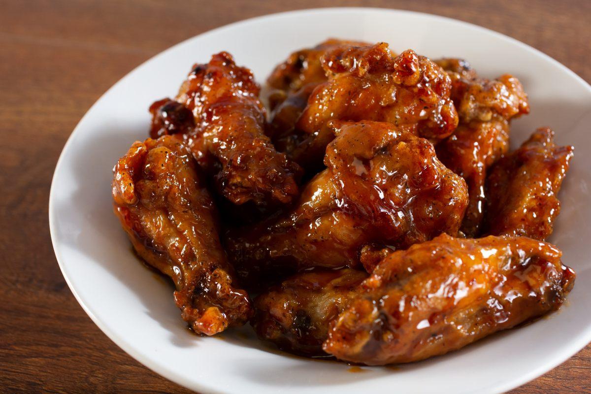 White bowl filled with glazed chicken wings coated in a thick, shiny, reddish-brown sauce, placed on a wooden surface.