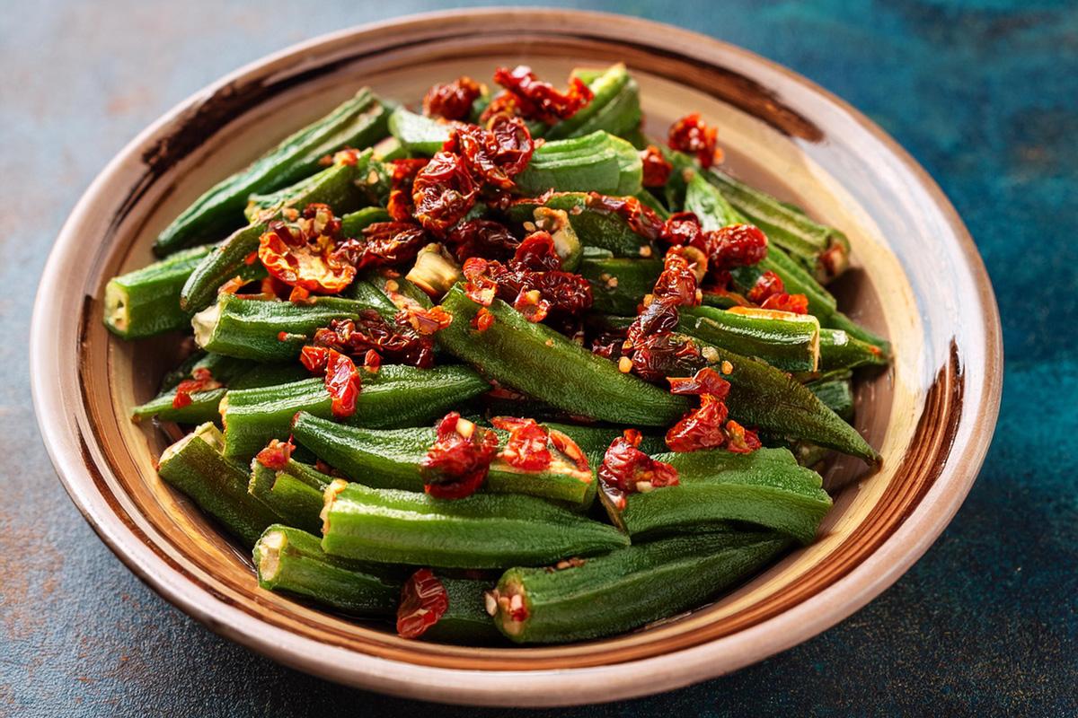 Colorful okra salad with sliced fresh green okra, sun-dried tomatoes, and spices served in a decorative bowl on a textured blue surface - GAI.