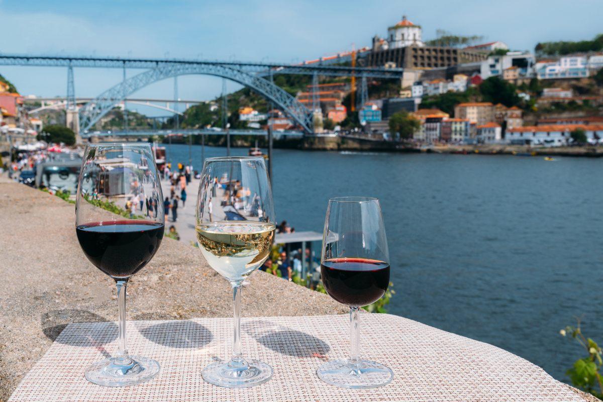 Three glasses of Protuguese wine, red, white, and dark red, on a table overlooking a scenic riverside Proto town with its bridge and colorful buildings on a sunny day.