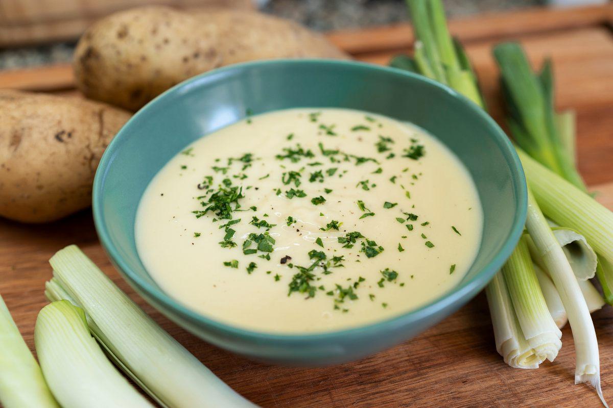 Close-up of a bowl of creamy vichyssoise soup garnished with fresh herbs, placed on a wooden board with raw leeks and potatoes in the background.