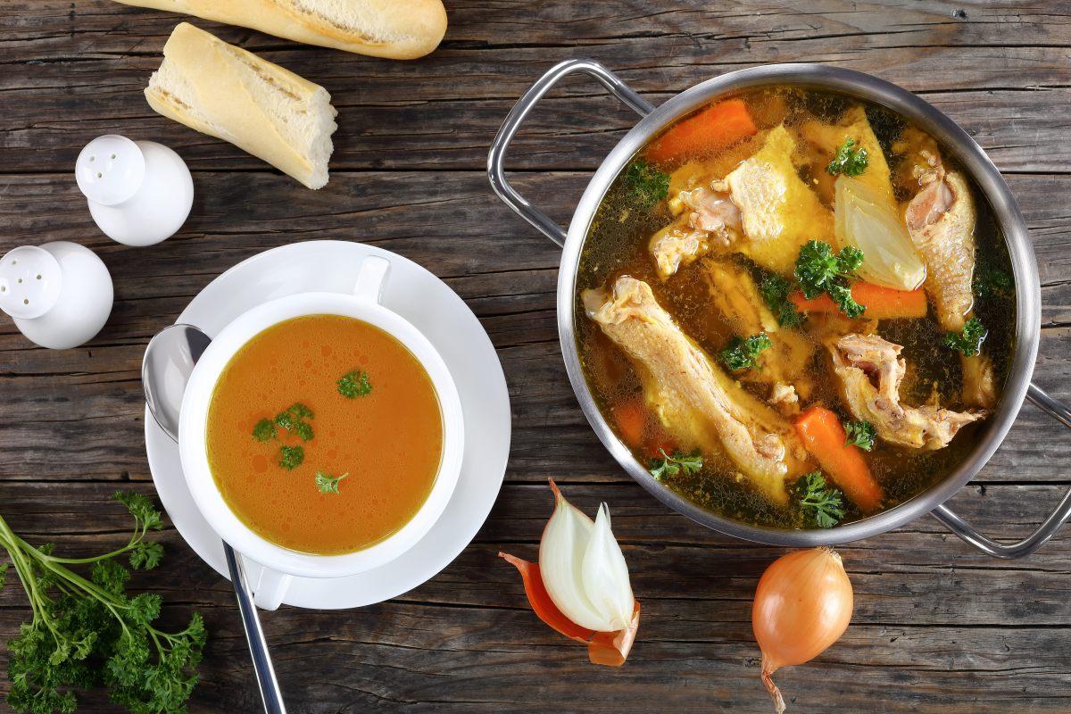 Clear chicken broth in a bowl. A pot with the rest of the broth with the ingredients is on the side.