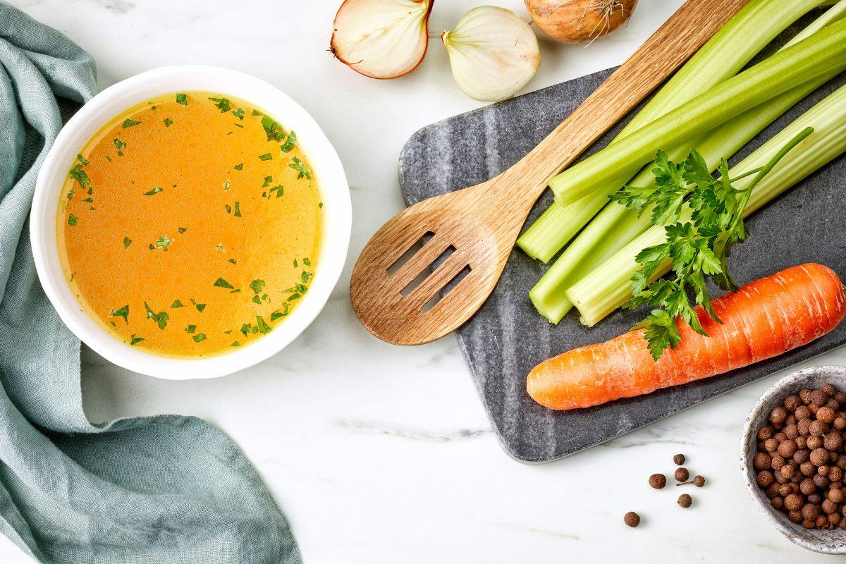 Bowl of chicken broth on a table, with some of the fresh ingredients by the side.