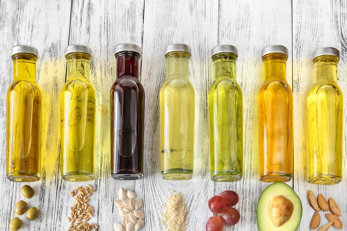 A row of different cooking oils in glass bottles with their source ingredients like olives, seeds, and avocado on a white wood background.