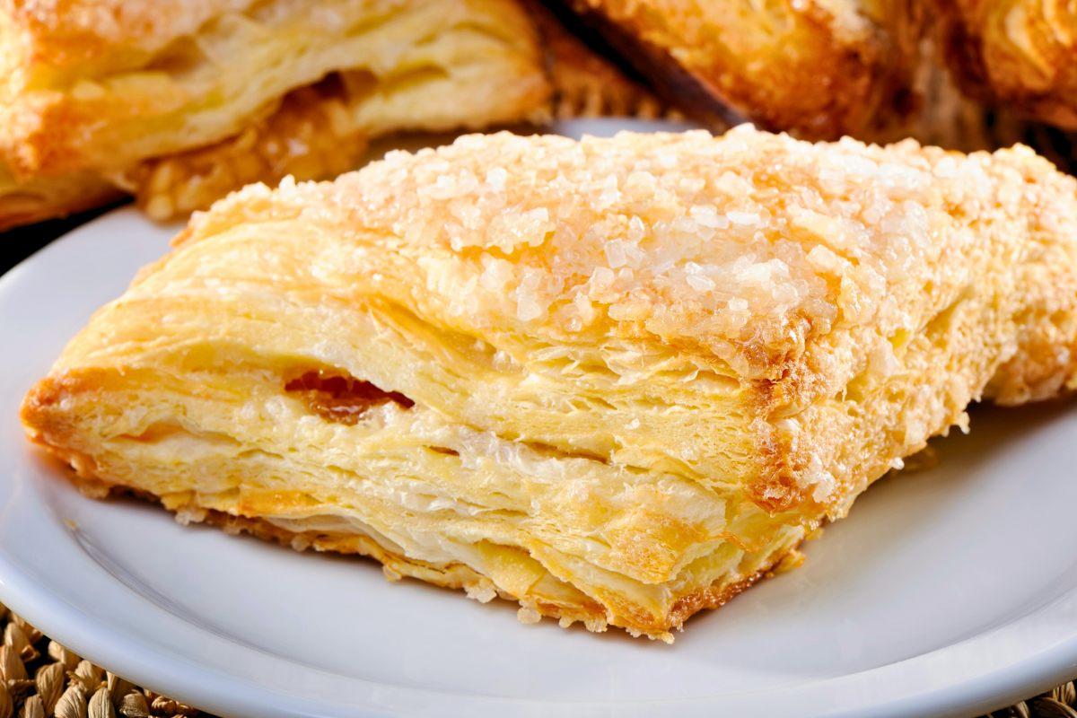 Close-up of a golden, flaky apple turnover on a plate, with warm apple-cinnamon filling peeking through the side and a light dusting of sugar.