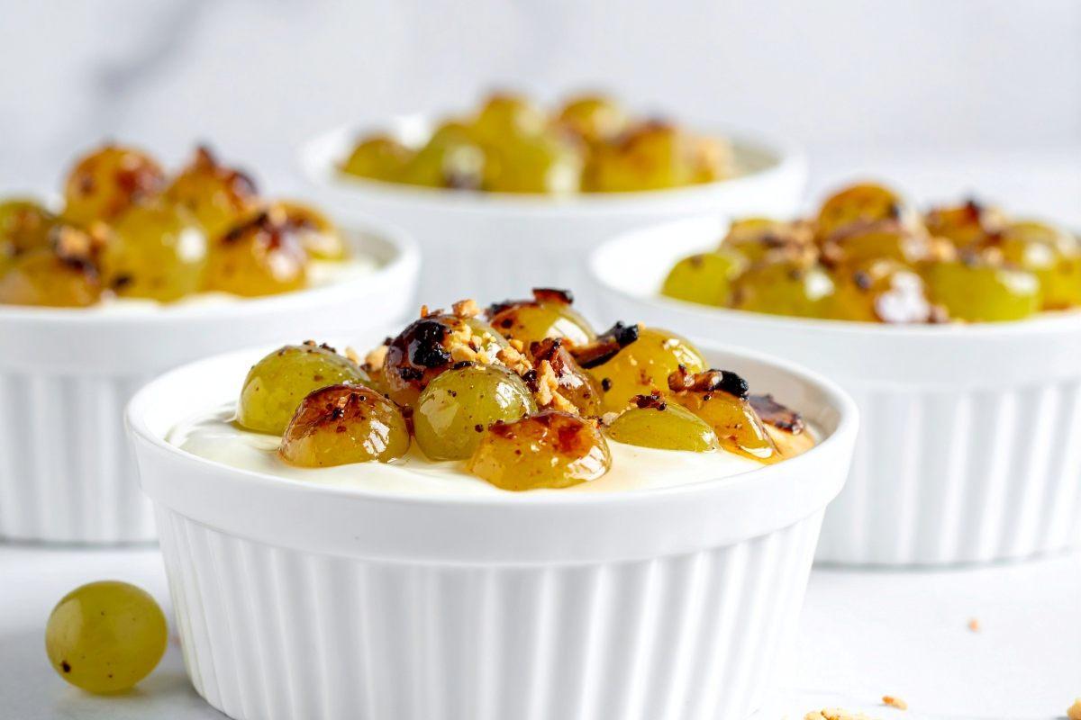 White ramekins filled with yogurt topped with roasted green grapes and toasted nuts on a bright white background.