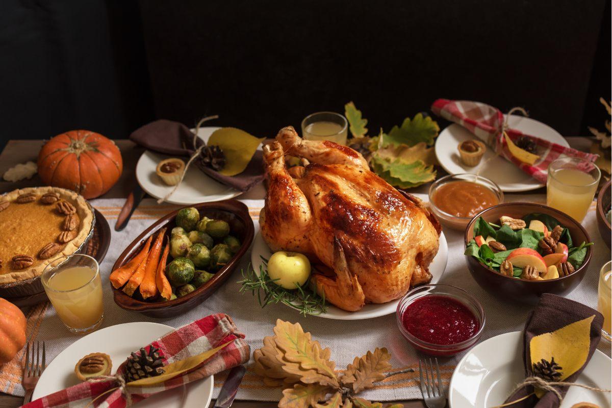 Holiday dinner table with a roasted turkey in the center, pumpkin pie, vegetables, salad, cranberry sauce, and fall decorations.