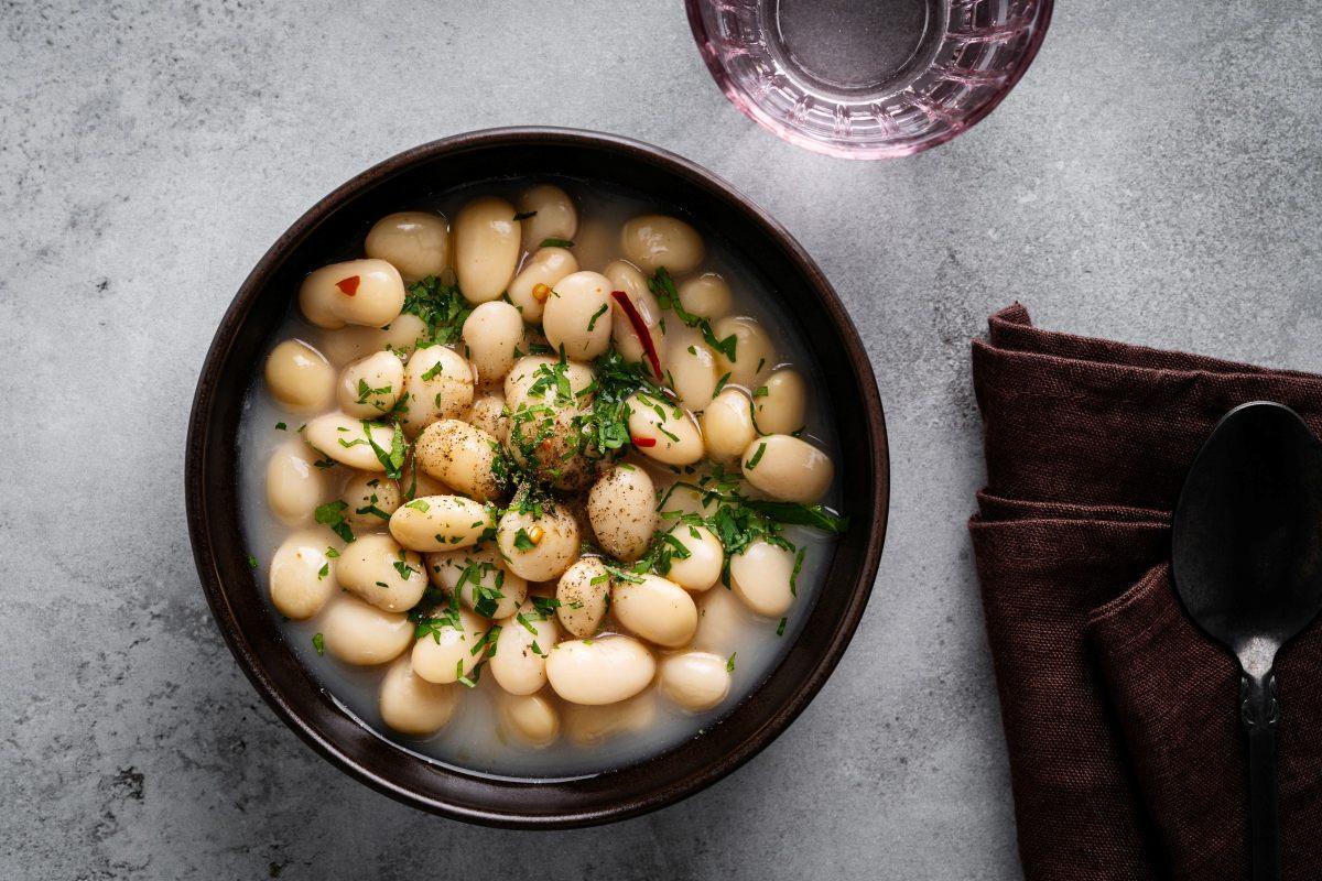 Bowl of white bean stew in broth, topped with fresh herbs, next to a brown napkin and spoon on a gray surface.