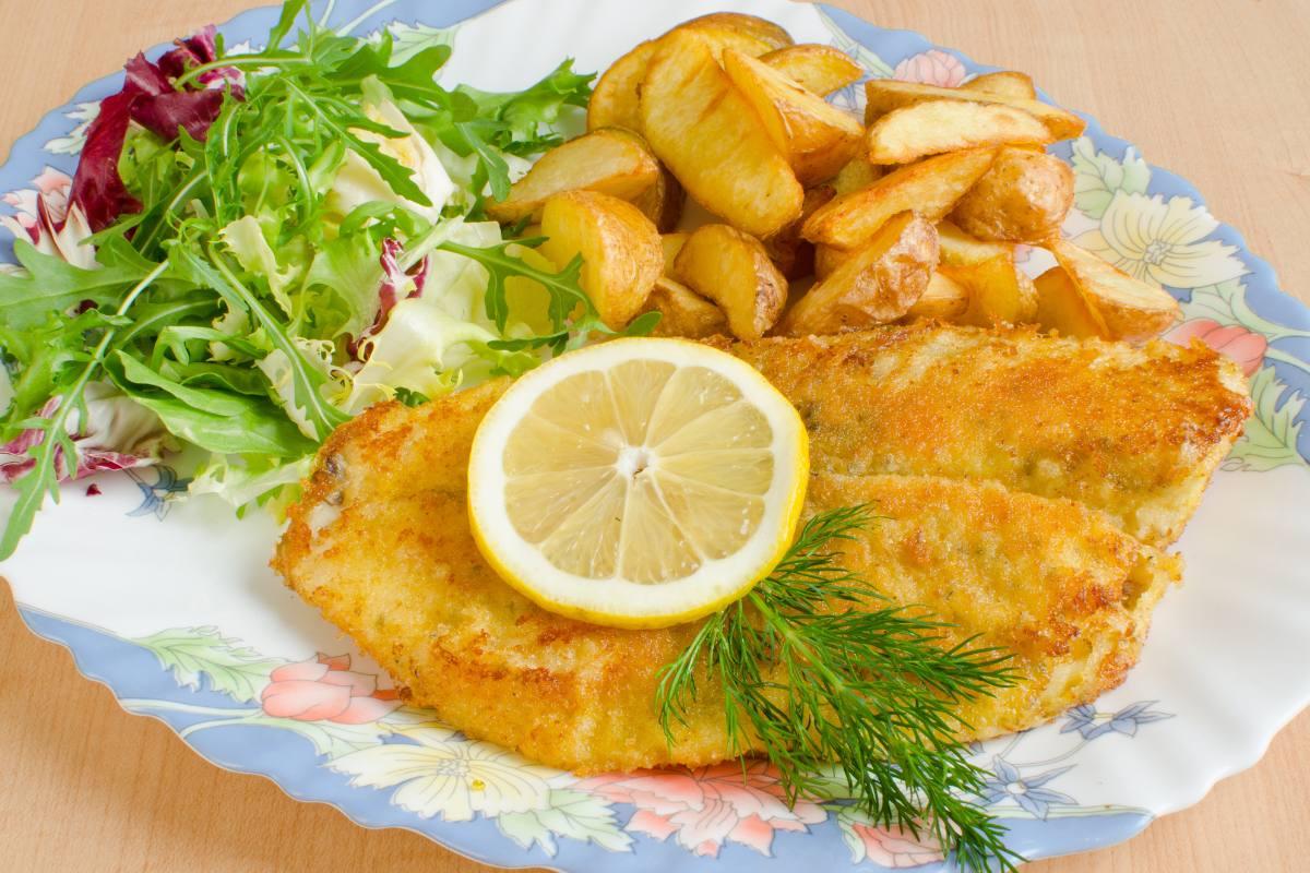 Crispy breaded tilapia fillet garnished with a lemon slice and dill, served with roasted potatoes and a mixed green salad on a floral plate.