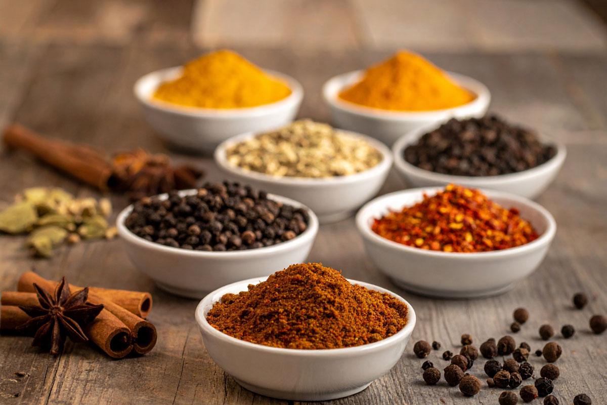 Small recipients with different ground spices like cumin, coriander, paprika, turmeric, cinnamon, black pepper, chili flakes, cardamom, garam masala and sumac. There are some spices on the table.