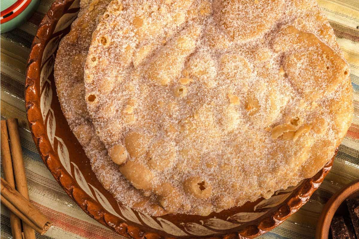 Sugar-coated buñuelos mexicanos on a rustic ceramic plate with cinnamon sticks.