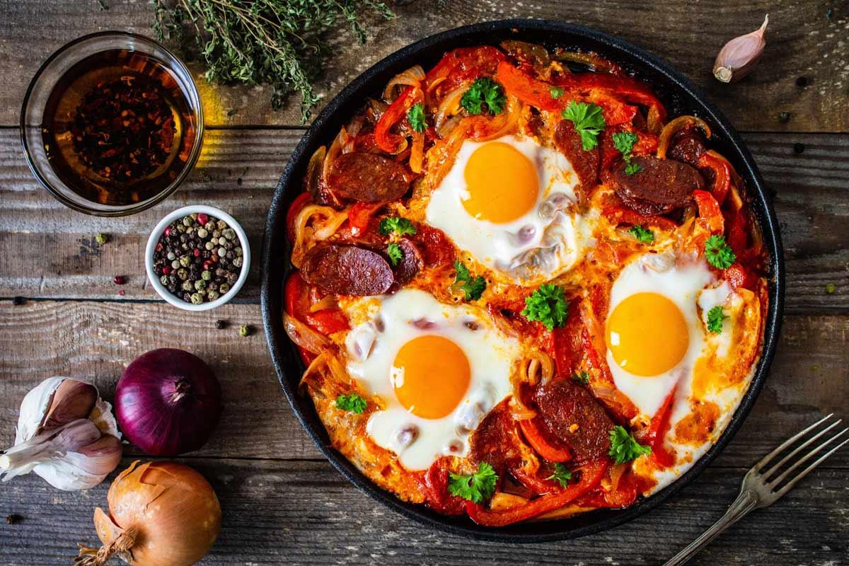 Chorizo shakshuka with four poached eggs, sliced cured sausage, and roasted red peppers in a tomato sauce, served in a cast-iron skillet on a rustic wooden table with garlic, onions, and spices.