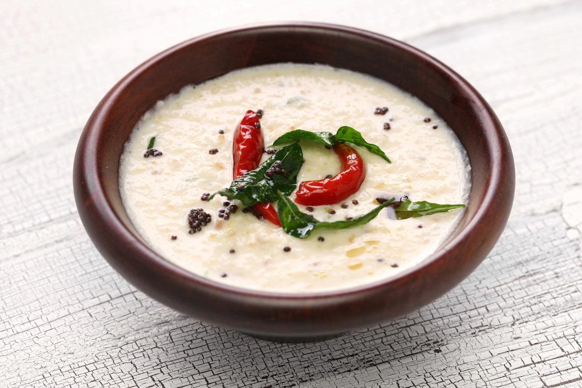 A clay bowl of smooth white coconut chutney garnished with a tempering of dried red chilies, curry leaves, and black mustard seeds, placed on a rustic white wooden surface.