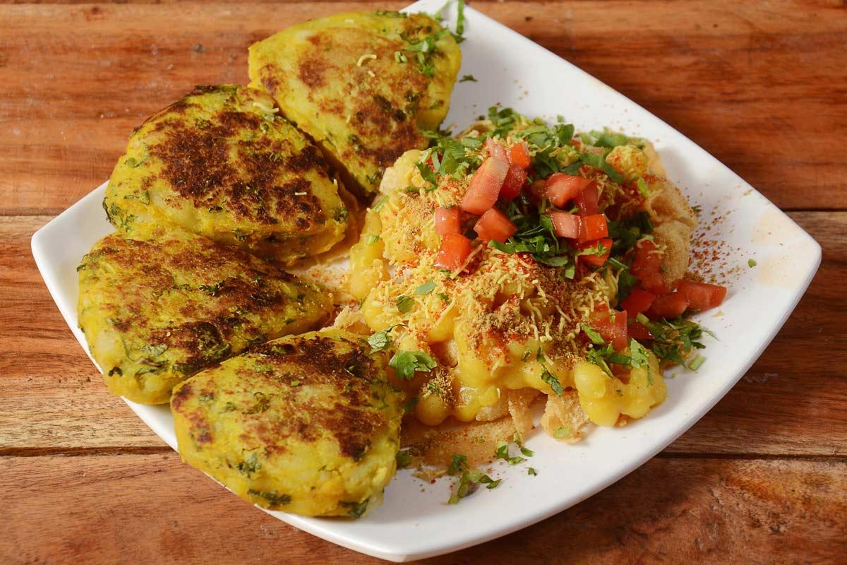 Aloo tikki chaat on a white plate: pan-fried spiced potato patties alongside chickpeas, diced tomato, sev, cilantro, and chaat masala, served on a wooden table.