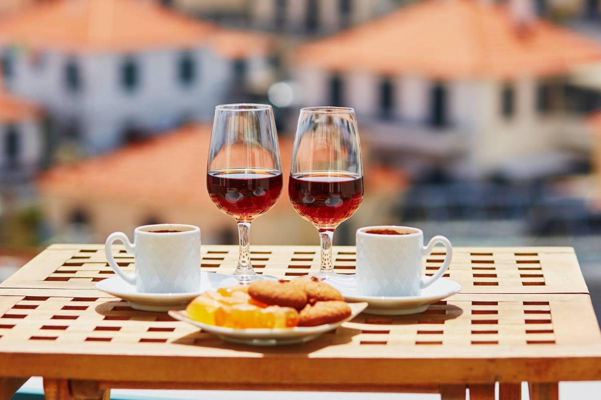 Two glasses of Madeira wine, two coffee cups, and a plate of sweets on an outdoor table overlooking blurred orange rooftops.