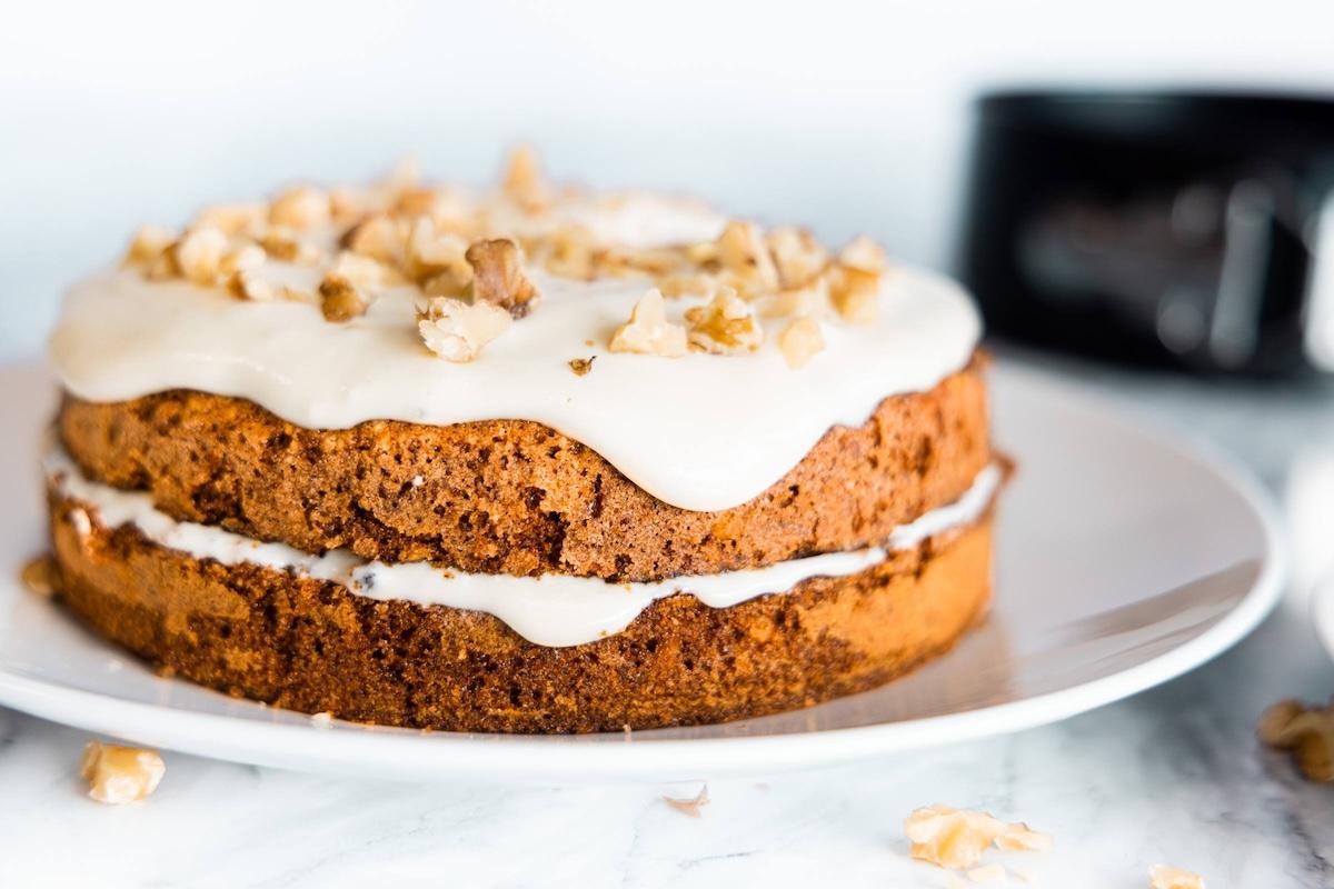 Two-layer carrot cake with cream cheese frosting and chopped walnuts on a white cake plate.