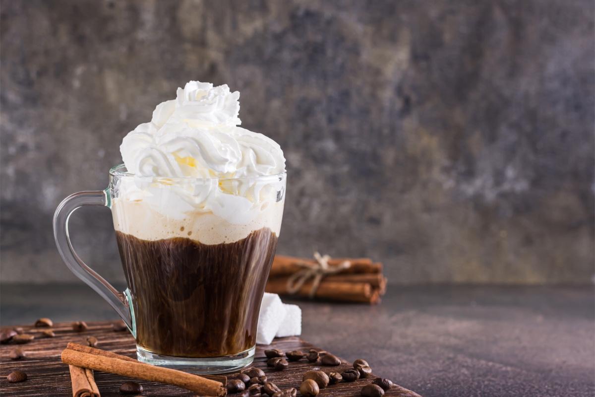 Glass mug of Einspänner coffee topped with whipped cream, surrounded by coffee beans and cinnamon sticks on a rustic table.