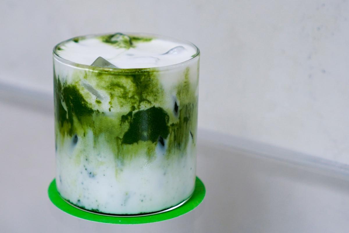 Glass of iced matcha Einspänner with green matcha swirls, milk, and ice cubes on a green coaster against a minimalist light background.