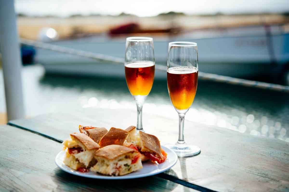 Two glasses of amber Marsala wine with a plate of savory Sicilian bites on a waterfront table.