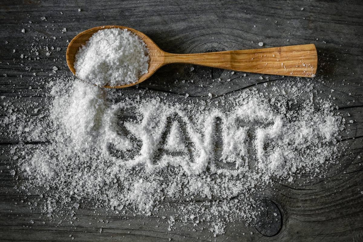 Wooden spoon filled with coarse salt on a dark table, with scattered salt spelling the word SALT.