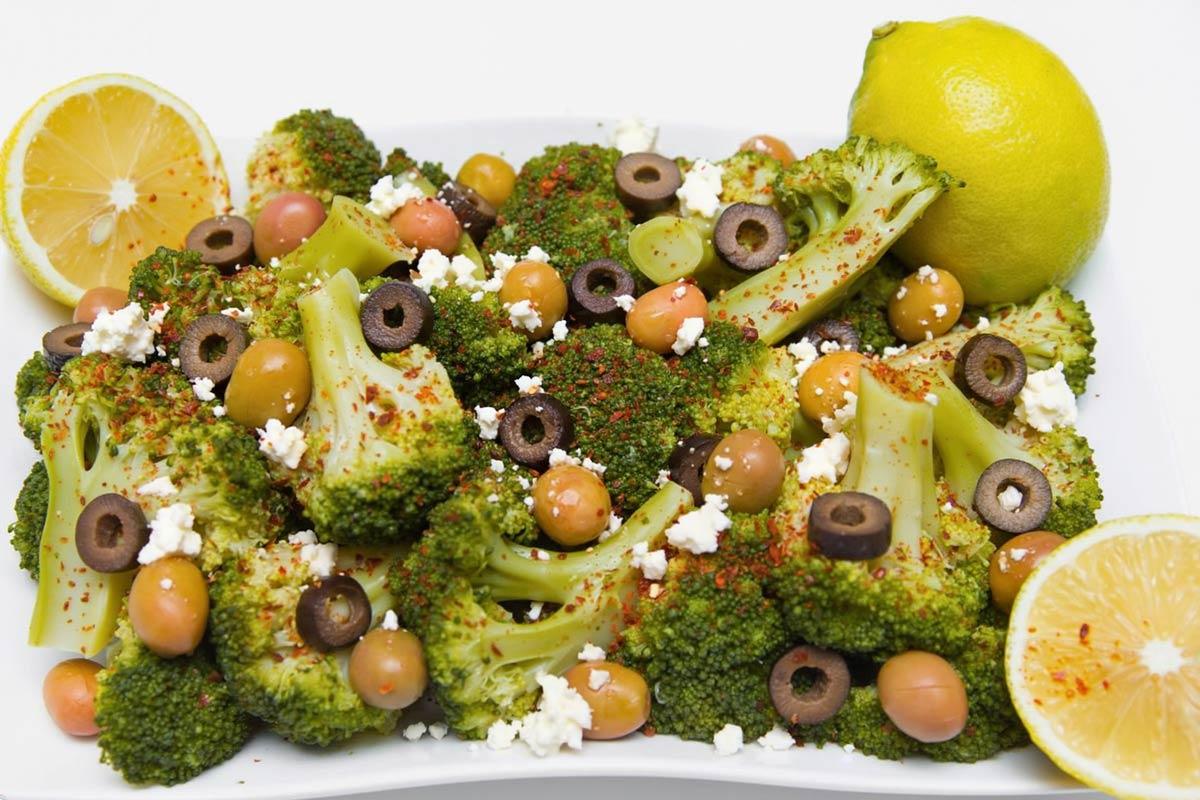 Broccoli salad with green and black olives, crumbled feta cheese, lemon, and red seasoning on a white platter.