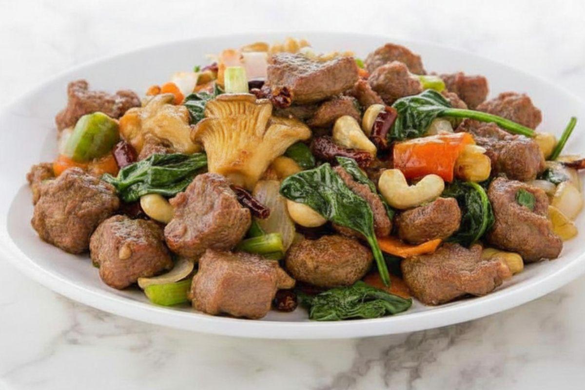 Cashew lamb stir-fry with vegetables, spinach, mushrooms, and dried chilies on a white plate.