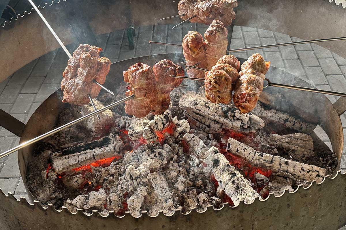 Several rolled meat roasts on metal spits cooking over glowing embers and burning logs in a large circular open-fire grill, with smoke rising from the coals.