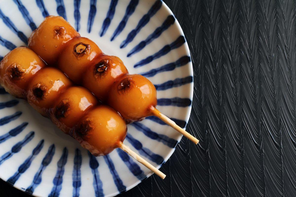 Two skewers of grilled Japanese dango rice dumplings coated in glossy sweet soy glaze, served on a white plate with blue striped decoration on a dark textured table.