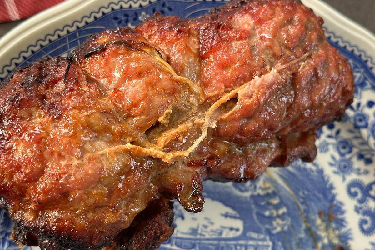 Close-up of a browned roasted meat roll with a crispy, glazed exterior, partially split open to reveal its juicy interior, served on a blue-and-white decorative plate.