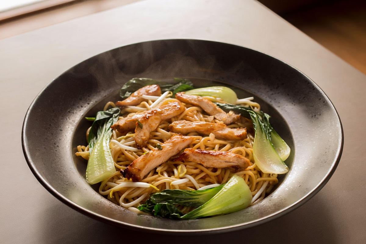 Steaming bowl of wok-fried noodles topped with sliced glazed chicken, bok choi, and bean sprouts. This is a synthetic image.