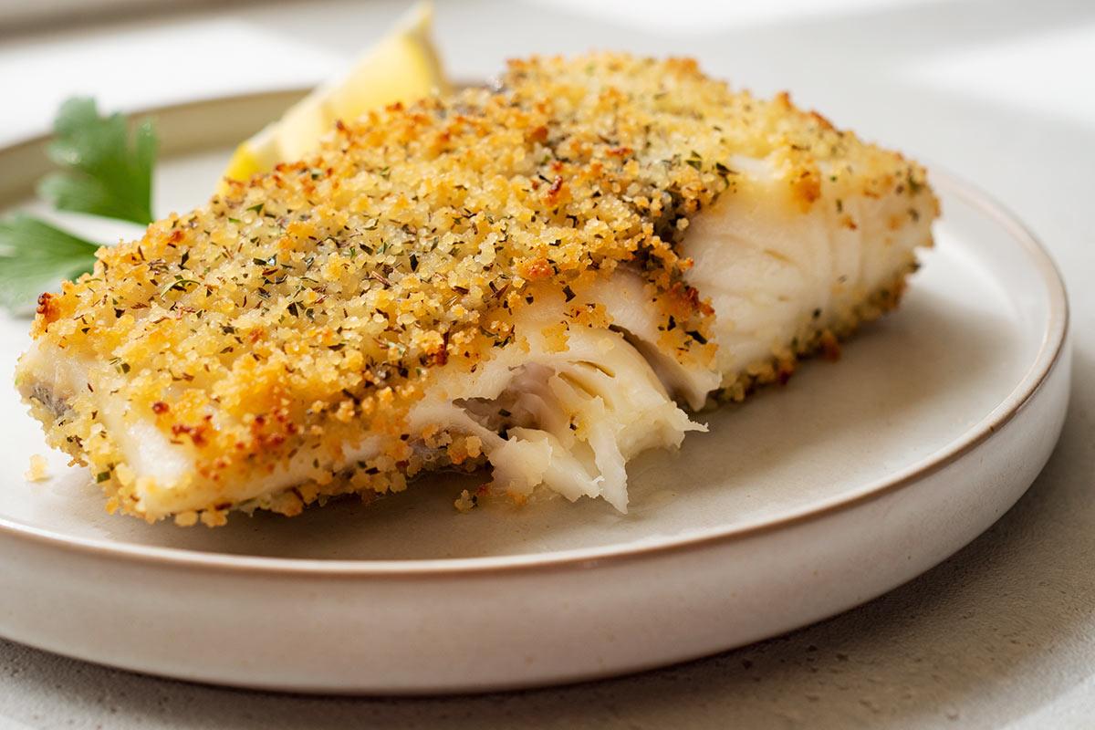 Parmesan-crusted fish fillet on a plate with golden breadcrumb topping, parsley, and a lemon wedge - synthetic image.