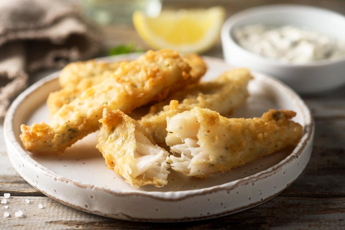 Golden beer-battered fish fingers on a white ceramic plate, one broken open to show the flaky white fish inside, with a lemon wedge and a bowl of tartar sauce in the background - synthetic image.