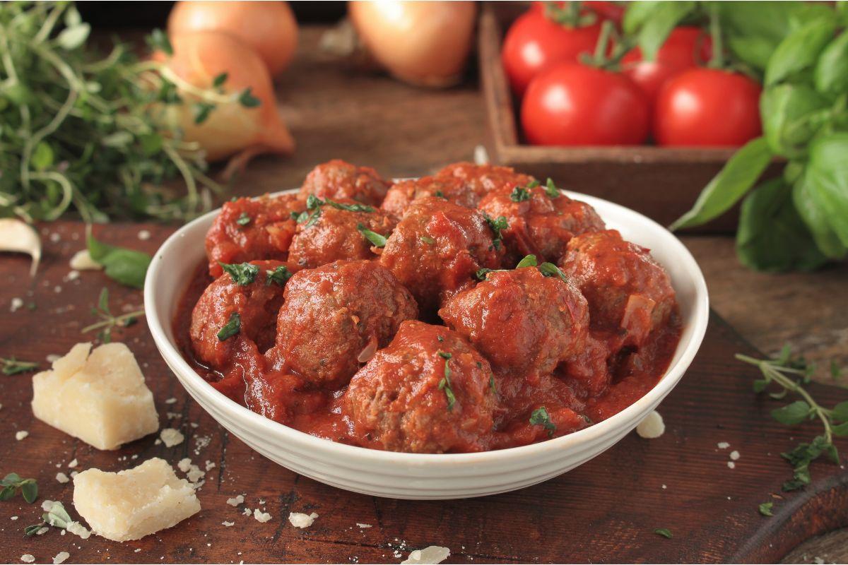 A white bowl of Italian meatballs in rich tomato sauce, sprinkled with fresh herbs, with basil, tomatoes, onions and chunks of Parmesan on a rustic wooden table.