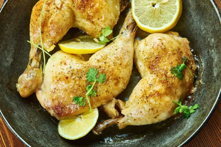 Close-up of four sumac-seasoned roasted chicken legs in a pan, garnished with fresh herbs and lemon wedges.