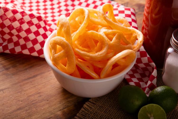White bowl filled with crispy, golden, ring-shaped snacks on a wooden surface. Behind the bowl is a red and white checkered cloth. A glass container with a dark liquid, a salt shaker, and two whole limes along with a lime half are also visible.