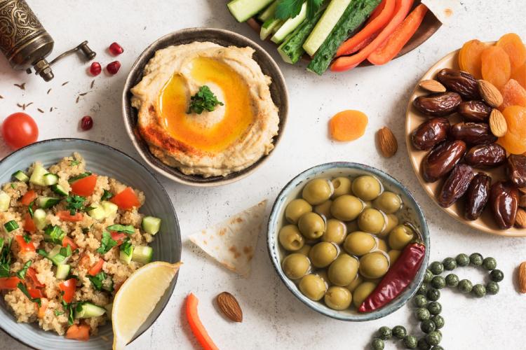 Middle Eastern food spread with a bowl of hummus topped with olive oil and paprika, fresh vegetable sticks, green olives with a red chili pepper, couscous salad with cucumber and tomato, and a plate of dried fruits with almonds. Garnishes include a vintage pepper grinder, cherry tomatoes, and a lemon wedge.