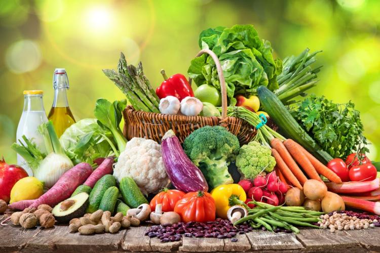 Colorful display of fresh organic vegetables and fruits, including bell peppers, carrots, lettuce, broccoli, and tomatoes, on a wooden table outdoors.