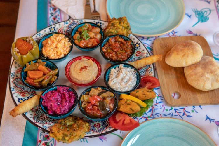 Top view of a Turkish mezze platter with assorted vegetable dishes, hummus, stuffed grape leaves, bread rolls, and dips served in small bowls on a patterned tablecloth.