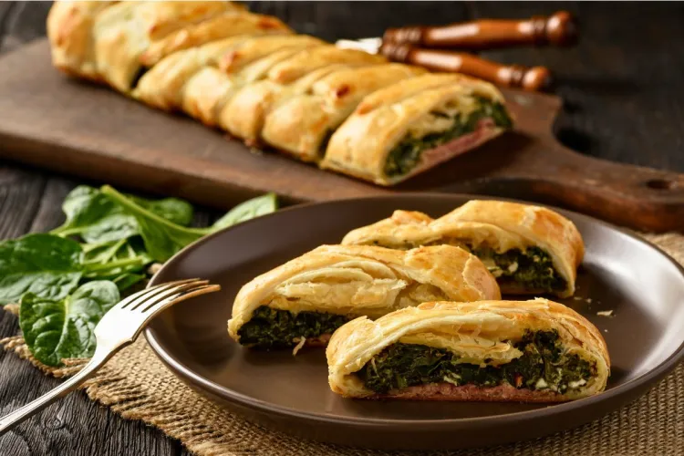 Close-up of a puff pastry pie filled with spinach, ham, and cheese, sliced on a dark brown plate with fresh spinach leaves on the table and the remaining pie on a wooden cutting board in the background.