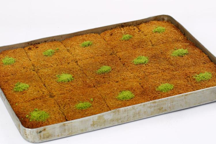 A rectangular baking tray filled with baked kataifi cut into squares, each topped with chopped pistachios.