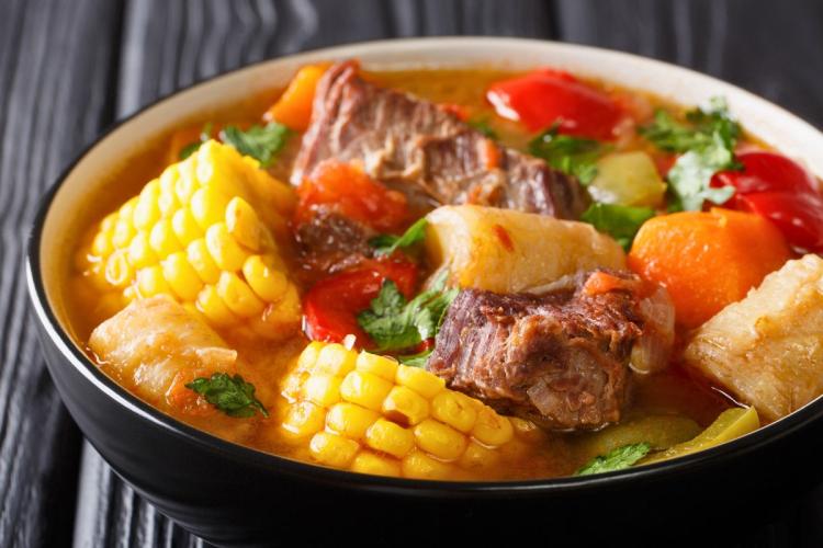 A bowl of sancocho, a traditional Latin American stew, featuring beef, corn on the cob, root vegetables, and fresh herbs in a golden broth.